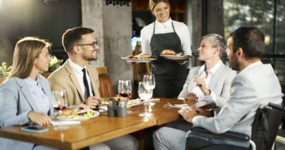 Diner sitting in a wheelchair with a group of other diners while a server brings their meals to the table.